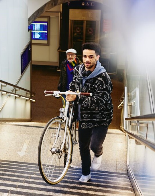 Man carrying a bike up the stairs in a train station. Other man walking up the stairs in the background.