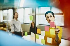 Two business women and a man in an office putting sticky notes on a window. Primary colors: blue.