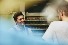 Two blue-collar workers talking to each other. Smiling. Tech environment. Caucasian man and man from India. Primary color blue.