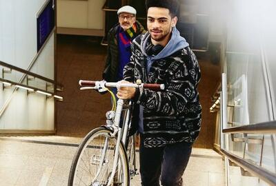 Man carrying a bike up the stairs in a train station. Other man walking up the stairs in the background.