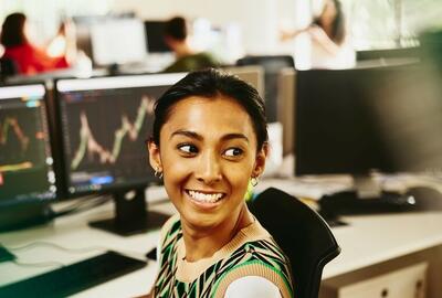 Smiling woman looking away from computer screens displaying financial information