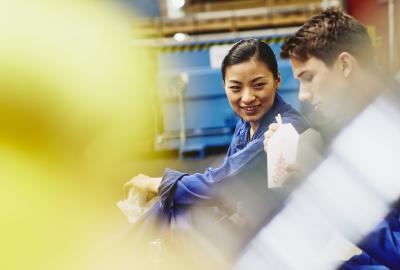 Male and female operator eating together. Food with chopsticks. Tech environment. Factory. Caucasian man and Asian woman. Primary color yellow. Secondary colors blue and white.
