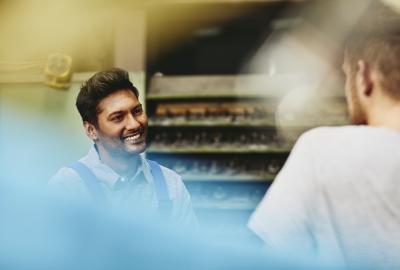 Two blue-collar workers talking to each other. Smiling. Tech environment. Caucasian man and man from India. Primary color blue.