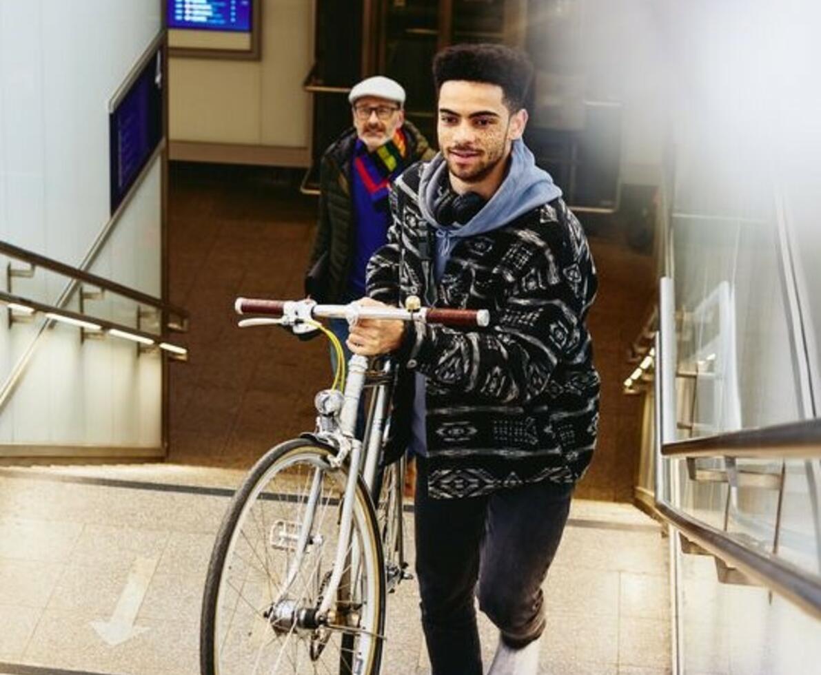 Man carrying a bike up the stairs in a train station. Other man walking up the stairs in the background.