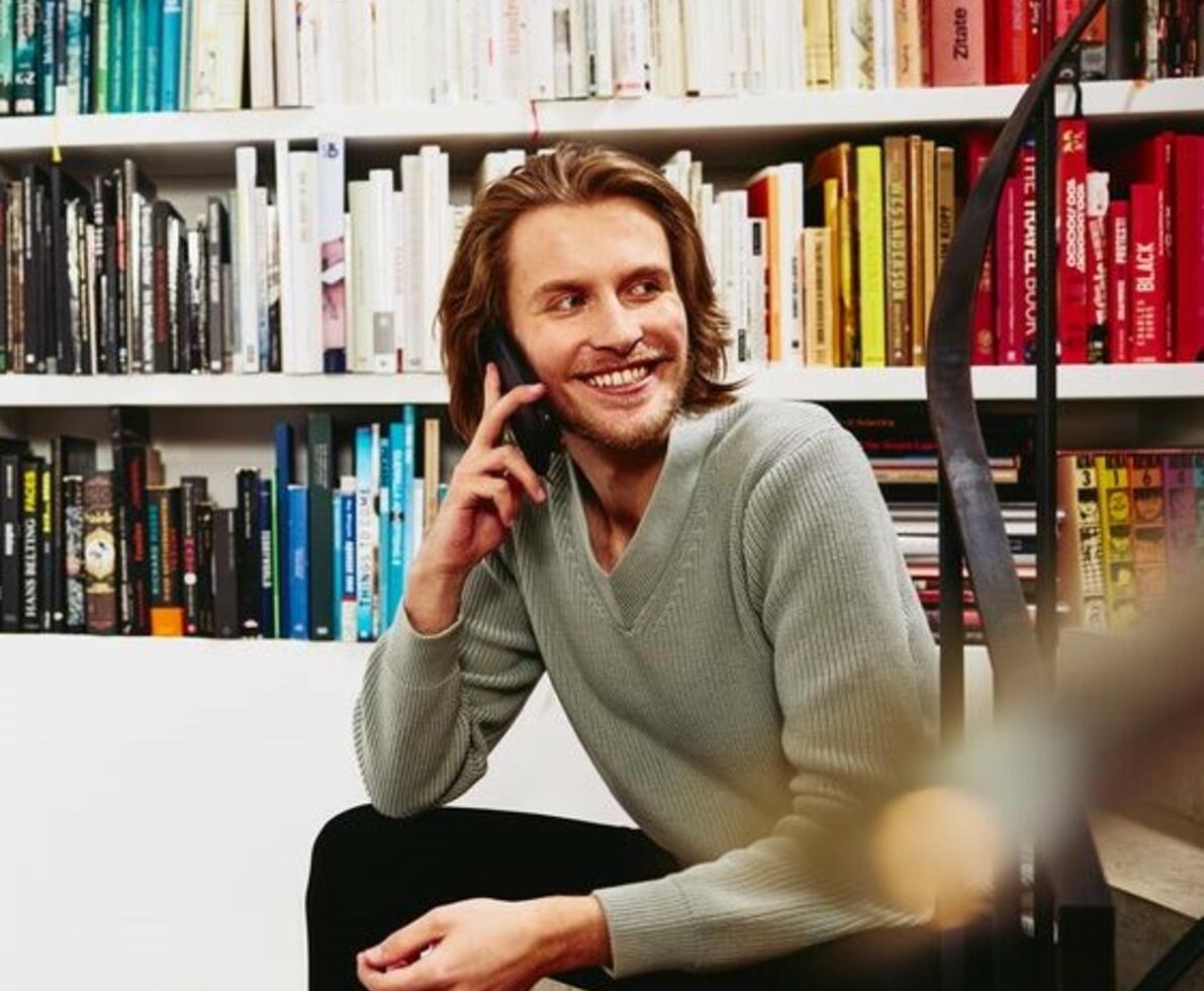 Smiling man sitting on stairs, having a phonecall. bookshelf in background