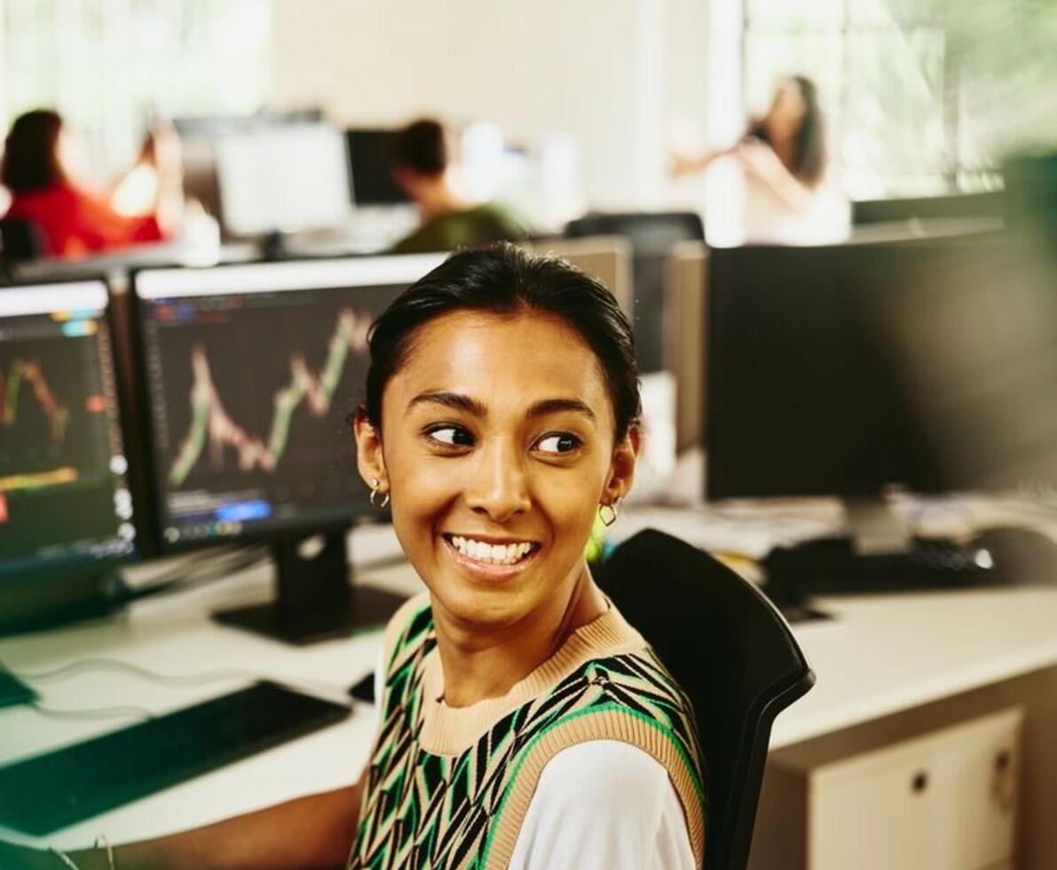 Smiling woman looking away from computer screens displaying financial information