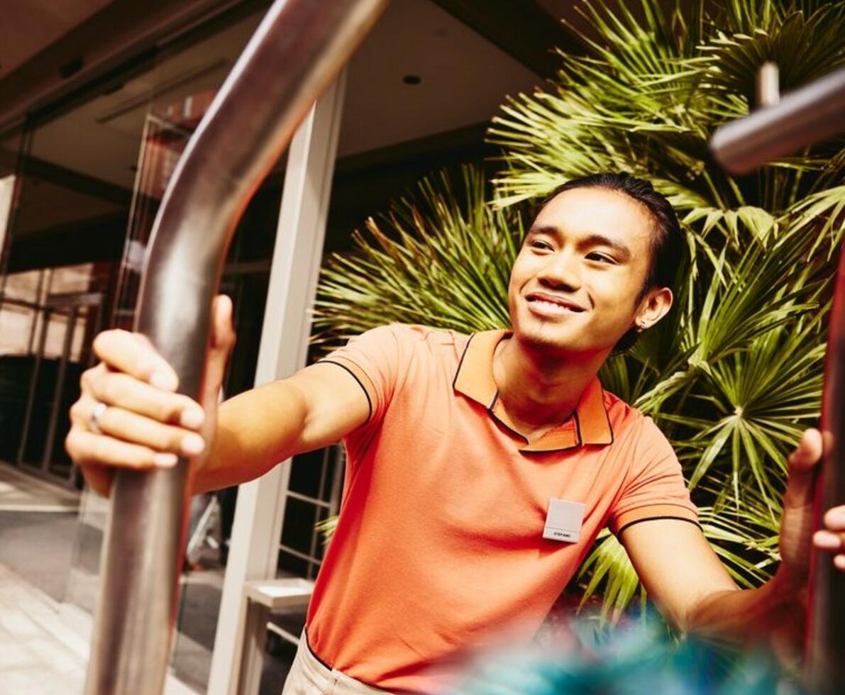 Smiling bellhop pushing a luggage trolley outside a hotel entrance.
