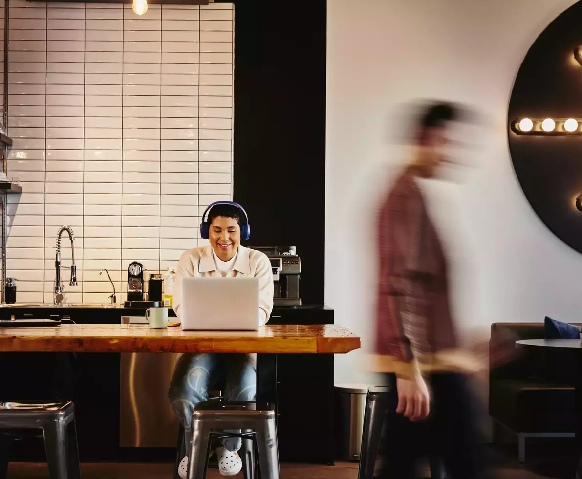 Man with headphones working on laptop at a table in a cafe while a man walks by