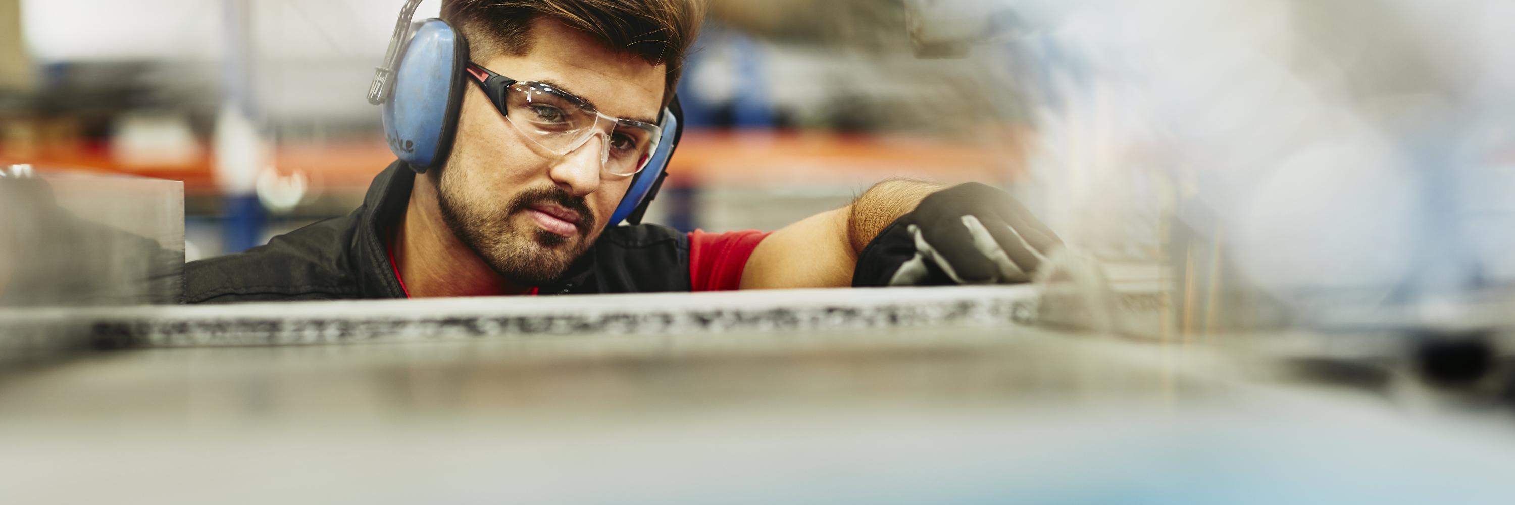 Male blue-collar worker in tech environment. Wearing protective ear covers, safety gloves. and safety goggles. Caucasian man. Groomed beard and mustache. Factory. Primary color blue. Secondary color cream.