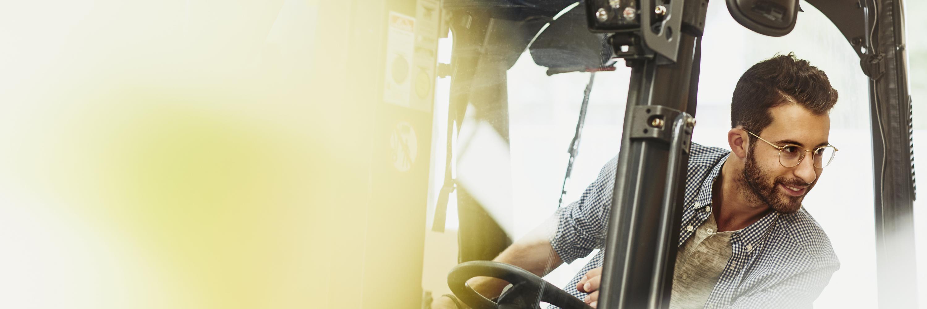 Male caucasian worker operating a forklift in a logistics environment. Looking to his left. Blue-collar. Smiling.  Wearing glasses. Groomed beard and moustache. Checkered shirt. Primary color cream.