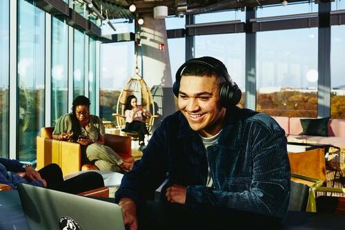 Smiling man with headphones and laptop sitting in a lounge environment.