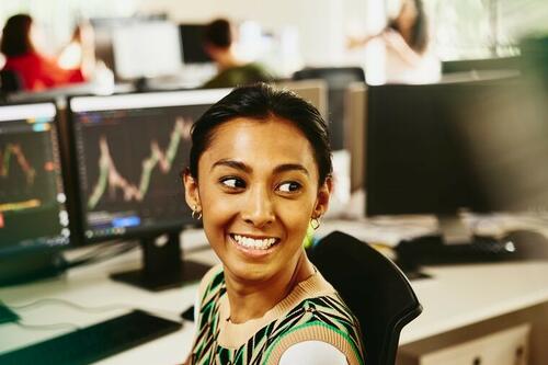 Smiling woman looking away from computer screens displaying financial information