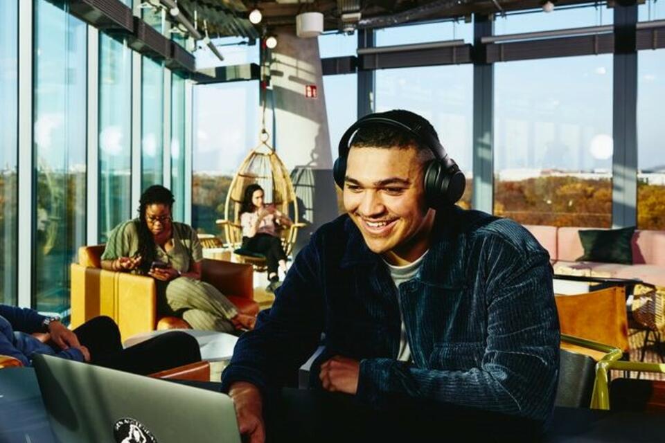 Smiling man with headphones and laptop sitting in a lounge environment.