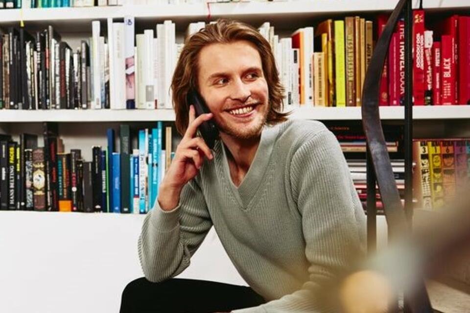 Smiling man sitting on stairs, having a phonecall. bookshelf in background