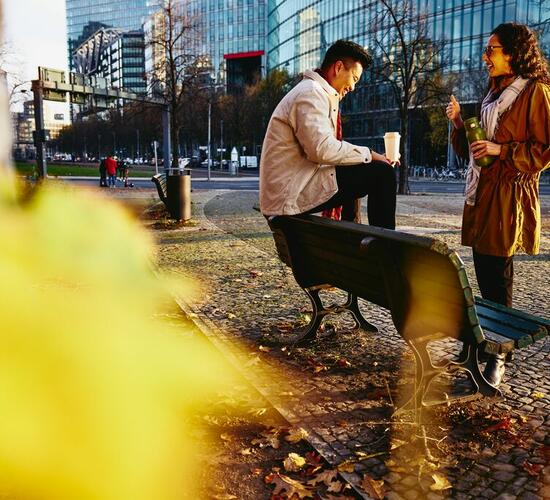 Man and woman holding drinks smiling and having a conversation. Man is sitting down on bench.