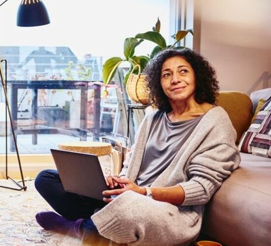 Woman leaning against a couch, sitting on the floor, with laptop on her lap