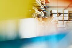 Male blue-collar worker operating a pallet loader in a logistics environment. Caucasion man. Neutral expression. Smooth-shaven chin. Moving parcels.  Primary colors yellow and blue. Secondary color cream.