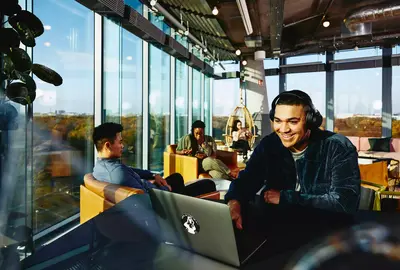 Smiling male with headphones and laptop sitting in a lounge environment.

