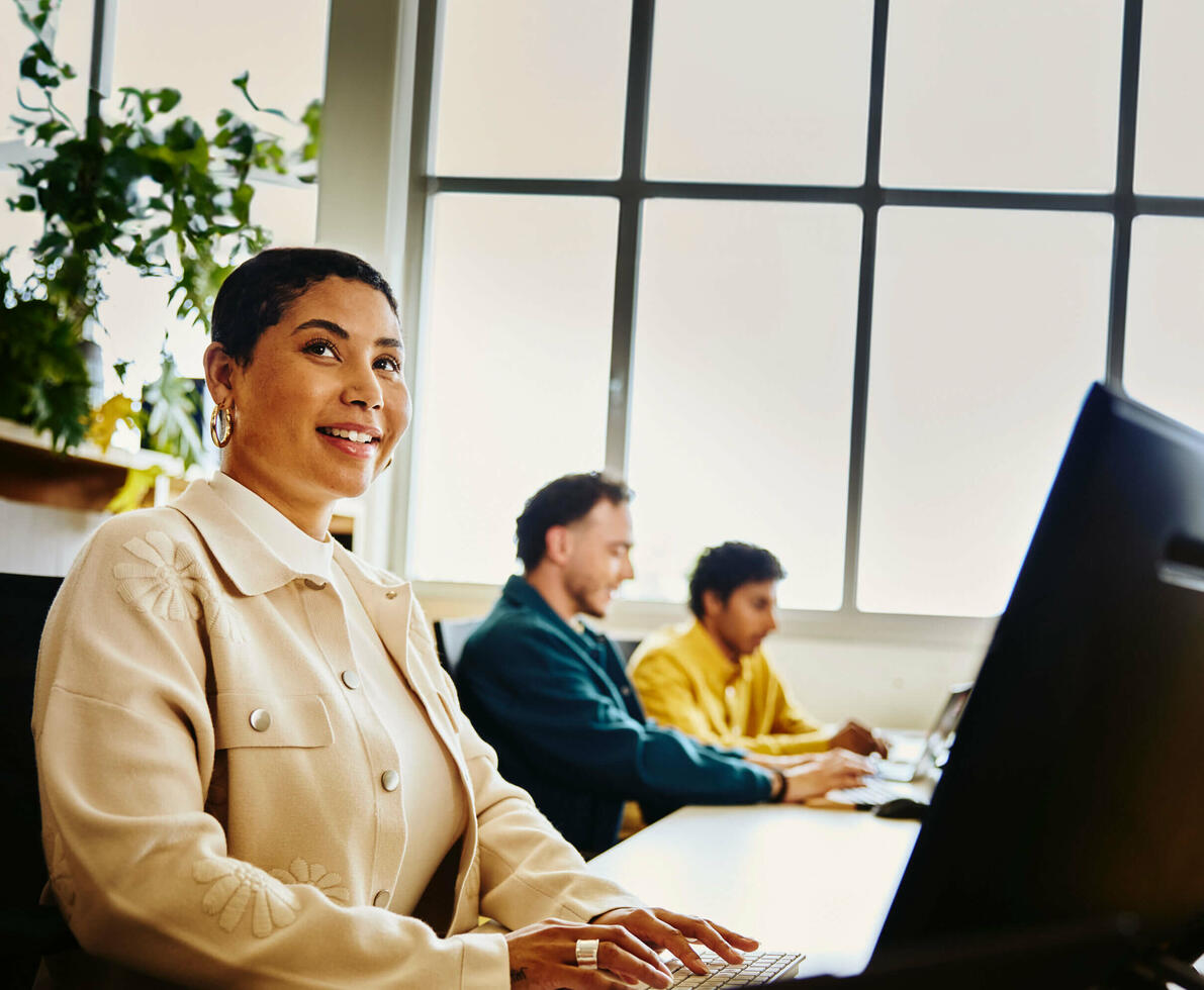 Female smiling, looking away from computer, two male sitting behind computer next to her