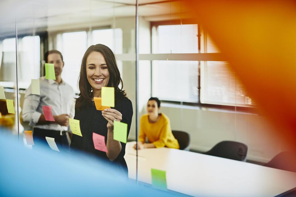 Two business women and a man in an office putting sticky notes on a window. Primary colors: yellow and blue.