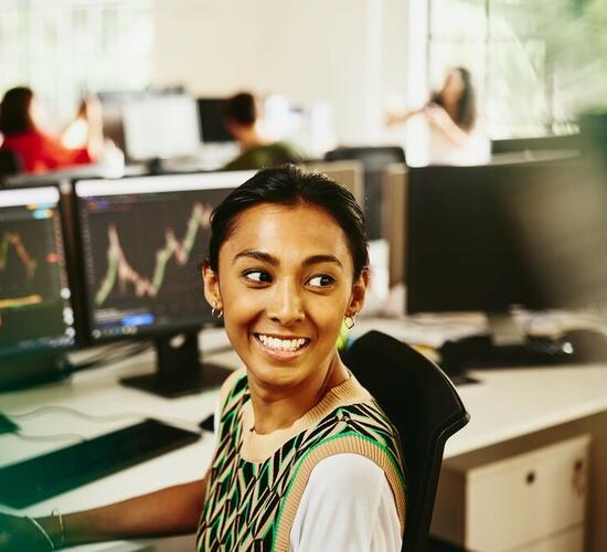 Smiling woman looking away from computer screens displaying financial information